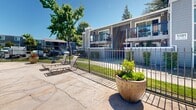 Located at 5109 Andrea Blvd in Sacramento, California, this outdoor amenity area serves as a central gathering space within a multi-building residential community. The centerpiece is a generously sized swimming pool with clear turquoise water, framed by a wide concrete pool deck that provides ample room for lounging and socializing. A separate circular hot tub/spa is positioned adjacent to the main pool, offering a relaxing retreat alongside additional seating. The pool area is enclosed by a sleek wrought iron fence with a gated entry, ensuring a secure and private environment for residents. Numerous lounge chairs and patio seating are arranged around the perimeter, ideal for enjoying Sacramento's sunny climate. Mature trees — including tall pines and deciduous varieties — surround the space, providing natural shade and a lush, park-like backdrop. Decorative planters with flowering plants and greenery add visual warmth to the patio. The multi-story residential buildings in gray and purple tones border the courtyard, with balconies overlooking the pool area. The open-sky setting allows for abundant natural light, making this an inviting outdoor destination throughout the day.