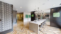 Located at 3120 Smith St in Houston's Montrose neighborhood, this thoughtfully designed business lounge and co-working space offers a sophisticated environment across a single floor. The space is unified by a striking chevron-patterned hardwood floor that runs throughout, lending warmth and visual continuity to every area.

The central hub features a long communal work table anchored beneath a dramatic multi-globe branching chandelier, creating an eye-catching focal point. Bold artwork, including vibrant green malachite-inspired panels and a large-scale tree ring mural, adds curated artistic character to the walls. A glass-enclosed private conference room with patterned carpet and ergonomic seating provides a quiet setting for meetings, accessible via sliding glass doors.

Compact individual workstations are built into dark-framed alcoves lined with textured geometric wallpaper, each equipped with a monitor and dedicated desk — ideal for focused, private work. A built-in refreshment station with a coffee machine and mini-refrigerator is conveniently integrated nearby. A cozy banquette nook with patterned upholstery and laser-cut decorative screens offers an additional informal meeting or lounge area. The space balances open collaboration with private productivity, all within a cohesive, design-forward aesthetic.