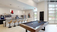 Located at 100 Pine St W in Wilson, NC, this well-appointed community amenity space offers a thoughtfully designed layout across a single generous floor. The open-concept living area serves as the heart of the space, featuring striking herringbone-patterned light wood flooring, soaring ceilings, and a bold mauve accent wall adorned with decorative gold leaf wall art. Large glass partition walls and oversized garage-style windows flood the interior with abundant natural light, creating an airy and welcoming atmosphere. The living area is furnished with a sectional sofa, accent chairs, a patterned area rug, and a wall-mounted television, making it ideal for relaxed gatherings. A pool table and additional game and dining tables provide further entertainment options. The fully equipped kitchen features navy cabinetry with gold hardware, a large central island with quartz countertops, stainless steel appliances including a full-size refrigerator and range, an eye-catching chevron tile backsplash, and open floating shelves. Pendant lighting adds a modern touch. A separate conference room offers a professional setting with long white tables, ample seating, a whiteboard, and a contemporary circular chandelier overhead, making it well-suited for meetings or events. The space blends modern design with functional versatility throughout.