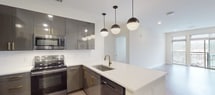 Located at 3718 University Dr in Durham, NC, this single-floor unit showcases a contemporary design with carefully selected finishes throughout. The kitchen serves as a central highlight, featuring high-gloss dark cabinetry accented with gold hardware, a full suite of stainless steel appliances, and a crisp white subway tile backsplash. A generous peninsula island with a white quartz countertop and undermount sink fitted with a brushed gold faucet provides additional workspace and casual seating. Globe pendant lights suspended overhead add a modern, stylish touch.

Light wood-style flooring extends through the kitchen and main living areas, creating a warm, cohesive aesthetic. The bedrooms are carpeted for comfort and each include a ceiling fan with recessed lighting, as well as windows fitted with blinds that allow natural light. The bathrooms are finished with granite countertops, dark-framed cabinetry, matte black fixtures, and bold framed mirrors. Each bathroom is positioned adjacent to a walk-in closet with wire shelving, offering practical storage. The overall ambiance is clean, modern, and move-in ready.