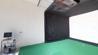This single-floor commercial space is purpose-built for indoor golf simulation, combining a functional hitting bay with a welcoming reception area. The main entry area is characterized by soaring ceilings, exposed ductwork, and a large glass storefront door flanked by floor-to-ceiling windows that flood the polished concrete floors with abundant natural light, offering clear views of the surrounding streetscape. A ceiling-mounted projector is positioned centrally overhead, serving as the technological centerpiece of the simulator setup. Two armchairs and a computer workstation are arranged near the entrance, creating a casual waiting and control area for guests. The simulator bay itself is enclosed by a structured black curtain system that defines the hitting zone and minimizes light interference, ensuring optimal screen visibility. Green artificial turf lines the hitting area, complete with a tee pad and a large white projection screen at the rear. The enclosure effectively creates an immersive golf experience within the open room. Exposed HVAC components run along the ceiling, contributing to the utilitarian, modern commercial aesthetic of the overall space.