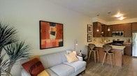 This single-floor space features an open-concept design that integrates the living area and kitchen into one cohesive environment. The living area is anchored by a light gray sofa adorned with decorative pillows, positioned beneath a bold, large-scale abstract painting in warm earth tones of orange, red, and brown, which serves as a striking focal point on the neutral white wall. A sleek arc floor lamp provides ambient lighting, complementing the space's understated elegance.

The kitchen area is accessible via a breakfast bar fitted with two stylish wooden bar stools with distinctive bentwood legs. The kitchen features dark wood cabinetry, light-colored countertops, and a pendant light that adds warmth to the cooking space. Framed artwork on the kitchen wall adds a curated, gallery-like feel that ties both areas together visually.

The flooring throughout consists of wood-look planks in a cool gray-brown tone, lending continuity and a contemporary feel across the space. The neutral wall palette allows the furnishings and artwork to stand out, creating a space that feels both functional and thoughtfully designed.