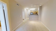 This single-floor residence offers a clean, open-concept layout that seamlessly connects the living area to the kitchen, creating a spacious and functional flow throughout. The living space is finished with plush wall-to-wall carpeting in a warm neutral tone, complemented by crisp white baseboards and smooth beige walls that lend a bright, welcoming ambiance. The kitchen features light wood cabinetry, dark granite-style countertops, and a raised breakfast bar peninsula that separates it from the main living area. Stainless steel appliances and a built-in microwave add a modern, practical touch to the cooking space, which is illuminated by a ceiling-mounted fluorescent light fixture. Track lighting installed along the kitchen-living transition ceiling provides additional directed illumination, enhancing both function and style. A hallway visible from the main living area leads to additional rooms, with white panel doors offering clean architectural continuity. Ceiling-mounted vents ensure efficient climate control throughout. The overall aesthetic is neutral, tidy, and move-in ready, with a cohesive color palette of beige, white, and light wood tones that creates a calm, comfortable atmosphere suited to a variety of interior styles.