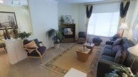 This single-floor space centers around a thoughtfully arranged living area that balances comfort and elegance. The room features a corner fireplace unit set within a wooden media cabinet, serving as a natural focal point and adding warmth to the space. Large windows dressed with layered drapes and blinds allow generous natural light to filter in, brightening the neutral, light-colored walls accented with crown molding.

The seating arrangement includes a mix of upholstered armchairs in deep blue and teal tones, complemented by a patterned area rug with earthy geometric borders that anchors the space. A pair of wooden side tables, one displaying a decorative tea service, adds a refined touch to the layout.

Lush indoor plants, including a large floor-standing palm, introduce vibrant greenery and a sense of life to the interior. Framed artwork and tasteful decorative accents adorn the surfaces, contributing to the curated aesthetic. The neutral carpeted flooring runs throughout, enhancing the warm, cohesive feel of the space. Overall, the room presents a well-appointed, comfortable environment with classic design elements and attention to decorative detail.
