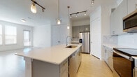 This single-floor residence showcases a contemporary design highlighted by a cohesive palette of white, gray, and light wood tones. The open-concept layout connects the kitchen and living areas seamlessly, creating a bright and airy atmosphere enhanced by natural light from multiple windows.

The kitchen serves as a focal point, centered around a generously sized island with a crisp white quartz countertop, an undermount sink, and brass-accented hardware. Gray flat-front cabinetry lines the perimeter walls, complemented by a marble-look tile backsplash and a full suite of stainless steel appliances, including a French door refrigerator, electric range, and over-the-range microwave. Brass pendant lights and adjustable track lighting add both functional and decorative illumination throughout.

The living area offers open, flexible space with light wood-tone flooring that extends throughout the residence. A hallway leads to a well-proportioned bedroom featuring multiple windows that bring in abundant natural light. The bathroom includes a dual-sink vanity with gray cabinetry and light countertops, a soaking tub, and a separate water closet. An additional bathroom features a similar modern vanity. A spacious walk-in closet with multi-level shelving and hanging rods provides ample storage.