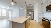 This single-floor residence showcases a contemporary design highlighted by a cohesive palette of white, gray, and light wood tones. The open-concept layout connects the kitchen and living areas seamlessly, creating a bright and airy atmosphere enhanced by natural light from multiple windows.

The kitchen serves as a focal point, centered around a generously sized island with a crisp white quartz countertop, an undermount sink, and brass-accented hardware. Gray flat-front cabinetry lines the perimeter walls, complemented by a marble-look tile backsplash and a full suite of stainless steel appliances, including a French door refrigerator, electric range, and over-the-range microwave. Brass pendant lights and adjustable track lighting add both functional and decorative illumination throughout.

The living area offers open, flexible space with light wood-tone flooring that extends throughout the residence. A hallway leads to a well-proportioned bedroom featuring multiple windows that bring in abundant natural light. The bathroom includes a dual-sink vanity with gray cabinetry and light countertops, a soaking tub, and a separate water closet. An additional bathroom features a similar modern vanity. A spacious walk-in closet with multi-level shelving and hanging rods provides ample storage.