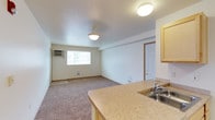 This single-floor space offers a practical and functional open-concept layout that seamlessly connects the kitchen and living area. The kitchen is equipped with a stainless steel sink set into a laminate countertop and features light wood cabinetry, providing ample storage. A breakfast bar-style counter separates the kitchen from the main living space, offering a convenient casual dining or workspace option.

The living area is finished with wall-to-wall carpeting that adds warmth underfoot. A window along the far wall allows natural light to filter in, brightening the room, while a wall-mounted air conditioning unit ensures climate comfort year-round. Pendant and flush-mount ceiling lights provide consistent overhead illumination throughout the space.

A white interior door leads to an additional room, offering flexibility for use as a bedroom or storage space. Neutral white walls throughout contribute to a clean, airy atmosphere, while standard baseboards and floor vents reflect a straightforward, well-maintained interior. The overall layout is efficient and well-suited for everyday living, with a logical flow between the kitchen and the open living zone.
