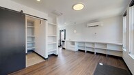 This single-floor unit is designed with an open, efficient layout that integrates living, sleeping, kitchen, and bathroom functions within one cohesive space. The main living area features warm wood-tone laminate flooring, white walls, and recessed lighting complemented by a flush-mount ceiling fixture, creating a clean and contemporary atmosphere.

A central built-in shelving unit and column divider help define separate zones within the open floor plan. The kitchenette is equipped with white cabinetry, a stainless steel refrigerator, a cooktop, and a countertop sink, with additional open shelving providing functional storage. A sliding barn-style door in dark-finished wood adds a modern design detail while providing access to the bathroom.

The bathroom features polished concrete flooring, a wall-mounted pedestal sink, a toilet, and a mirror, maintaining the unit's clean aesthetic. A separate sleeping area is bathed in natural light through a bank of three tall windows, with built-in shelving along the wall offering additional storage. A wall-mounted mini-split system provides climate control in both the main living area and sleeping zone. The overall design is minimal, functional, and modern throughout.