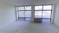 This single-floor space is defined by its striking wall of large, grid-paned windows that span nearly the full width of the main living area. The oversized windows flood the interior with natural light and provide unobstructed views of the surrounding neighborhood, including mature trees and adjacent rooftops. Light-colored, wall-to-wall carpeting covers the floor, contributing to a clean and neutral aesthetic throughout the space. The white walls and simple, unadorned ceiling create a minimalist backdrop that enhances the sense of openness. A through-wall PTAC heating and cooling unit is positioned beneath one of the window panels, ensuring year-round climate control. The corner window configuration allows light to enter from multiple directions, reducing the need for artificial lighting during daytime hours. Horizontal blinds on each window panel provide privacy and light management as needed. The overall layout is open and versatile, offering flexibility for various furniture arrangements. The combination of abundant natural light, neutral finishes, and a straightforward design makes this a practical and adaptable living space.