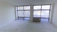 This single-floor space is defined by its striking wall of large, grid-paned windows that span nearly the full width of the main living area. The oversized windows flood the interior with natural light and provide unobstructed views of the surrounding neighborhood, including mature trees and adjacent rooftops. Light-colored, wall-to-wall carpeting covers the floor, contributing to a clean and neutral aesthetic throughout the space. The white walls and simple, unadorned ceiling create a minimalist backdrop that enhances the sense of openness. A through-wall PTAC heating and cooling unit is positioned beneath one of the window panels, ensuring year-round climate control. The corner window configuration allows light to enter from multiple directions, reducing the need for artificial lighting during daytime hours. Horizontal blinds on each window panel provide privacy and light management as needed. The overall layout is open and versatile, offering flexibility for various furniture arrangements. The combination of abundant natural light, neutral finishes, and a straightforward design makes this a practical and adaptable living space.