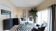This single-floor residence offers a thoughtfully designed living space that blends contemporary style with everyday comfort. The living area serves as the focal point of the home, featuring warm neutral-toned walls that create a welcoming and versatile backdrop for the bold, modern furnishings throughout. A plush white sofa adorned with black and white patterned accent pillows anchors the seating arrangement, complemented by a sleek glass-topped coffee table and a dark upholstered armchair. A striking black and white geometric area rug defines the space and adds a dynamic visual element underfoot. Two decorative mirrored wall panels reflect light and contribute to the room's contemporary aesthetic, while a tall, lush indoor palm plant introduces a natural, organic element that softens the overall design. Large sliding glass doors allow an abundance of natural light to flood the interior and provide a seamless transition to an outdoor area, offering views of the surrounding greenery. The textured ceiling adds subtle architectural character, and the neutral carpeting throughout ties the space together cohesively. The overall ambiance is modern, curated, and inviting, balancing bold design choices with a clean, airy atmosphere.