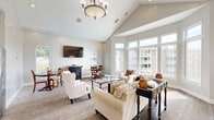 Located at 4085 Warner Ave in Hyattsville, Maryland, this single-floor combined living and dining space offers a thoughtfully designed, light-filled interior that maximizes both comfort and functionality. The most striking architectural feature is the dramatic vaulted ceiling, which rises steeply to create a sense of openness well beyond the room's footprint. Floor-to-ceiling windows wrap around the living area, flooding the space with abundant natural light and offering sweeping views of an outdoor pool, manicured lawn, and neighboring buildings. The neutral gray and white color palette throughout the walls and trim enhances the airy, welcoming ambiance. The living area is anchored by plush seating, a glass-top coffee table, and a wall-mounted flat-screen television, creating an inviting setting for relaxation. Transitioning seamlessly from the living area, the dining space accommodates a classic wooden round table and chairs, positioned near a window that frames lush outdoor greenery. A wall-mounted television and a decorative side cabinet add both style and practicality to the dining nook. Carpeted flooring in a woven neutral pattern ties the two areas together cohesively. Recessed lighting complements the natural illumination, ensuring the space remains bright and functional at all hours.