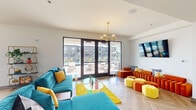 This single-level community room in Louisville, KY (40206) is a thoughtfully designed shared space that combines social, dining, and lounge functions within one open, flowing layout. Herringbone-patterned light wood flooring runs throughout, lending a cohesive, modern aesthetic to the entire space.

The lounge area is furnished with bold, jewel-toned seating—including a teal velvet sofa, richly textured orange sectional, and vibrant yellow ottoman cubes—anchored by a statement chandelier with gold-toned globe pendants. A wall-mounted flat-screen television and colorful wall art add visual interest. Large black-framed French doors open directly to an outdoor pool terrace, flooding the space with natural light and providing seamless indoor-outdoor connectivity.

Adjacent to the lounge is a dedicated dining area featuring a natural wood table paired with mustard yellow upholstered chairs, illuminated by a matching globe chandelier. A gold-framed open shelving unit displays decorative objects and greenery nearby.

The kitchen and bar area is a standout feature, showcasing a teal-painted ceiling, geometric patterned tile backsplash, extended white quartz countertops, wood cabinetry, and a lengthy kitchen island with bar seating. Open shelving displays colorful books, plants, and décor, creating an inviting, café-like atmosphere. Additional windows provide natural light throughout.