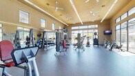 Located in Bella Terra in Richmond, TX, this well-appointed fitness center offers a generously sized, single-floor exercise space designed to support a full range of workout routines. The room features soaring ceilings fitted with multiple ceiling fans and sleek linear lighting, creating a bright and comfortable environment for exercise.

The space is comprehensively equipped with a variety of strength-training machines, free weights including a full dumbbell rack and kettlebell set, cable machines, and TRX suspension trainers. The cardio section includes multiple treadmills and upright bikes, positioned directly in front of expansive floor-to-ceiling windows that frame views of the outdoor pool and surrounding community — an inviting backdrop during any workout.

Full-length mirrors line one wall, enhancing the sense of openness and allowing users to monitor their form. The durable rubber flooring throughout provides safety and comfort underfoot. Wall-mounted flat-screen televisions are positioned throughout the space for entertainment. Natural light pours in from multiple window banks, including clerestory windows near the roofline, keeping the space feeling airy and energizing throughout the day. The warm neutral-toned walls and clean layout contribute to a welcoming, professional fitness environment.