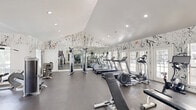 Located at 2000 Colony Park Dr in Birmingham, Alabama, this fitness center offers a well-appointed exercise space designed for a comprehensive workout experience. The room's most striking design element is its bold, abstract splatter-pattern wall treatment in black and white, which covers the walls from floor to ceiling and gives the space an energetic, artistic character that sets it apart from a typical gym environment.

The facility features vaulted ceilings with recessed lighting, contributing to a bright and open atmosphere. Large windows and glass-paned French doors line one wall, flooding the interior with natural light and providing views of the surrounding outdoor greenery. Full-length mirrors along one wall visually expand the space and support proper form during exercise.

The exercise room is comprehensively equipped for both cardiovascular and strength training. Cardio equipment includes multiple treadmills and elliptical machines, while the strength training area offers cable machines, multi-station weight equipment, free weights with a dedicated rack, a flat bench, and medicine balls. The durable rubberized flooring throughout supports safe, high-impact workouts. A wall-mounted television and a mounted hand sanitizer station are also present. The layout is thoughtfully organized to provide ample space between equipment zones, ensuring a comfortable and functional fitness experience.