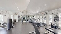 Located at 2000 Colony Park Dr in Birmingham, Alabama, this fitness center offers a well-appointed exercise space designed for a comprehensive workout experience. The room's most striking design element is its bold, abstract splatter-pattern wall treatment in black and white, which covers the walls from floor to ceiling and gives the space an energetic, artistic character that sets it apart from a typical gym environment.

The facility features vaulted ceilings with recessed lighting, contributing to a bright and open atmosphere. Large windows and glass-paned French doors line one wall, flooding the interior with natural light and providing views of the surrounding outdoor greenery. Full-length mirrors along one wall visually expand the space and support proper form during exercise.

The exercise room is comprehensively equipped for both cardiovascular and strength training. Cardio equipment includes multiple treadmills and elliptical machines, while the strength training area offers cable machines, multi-station weight equipment, free weights with a dedicated rack, a flat bench, and medicine balls. The durable rubberized flooring throughout supports safe, high-impact workouts. A wall-mounted television and a mounted hand sanitizer station are also present. The layout is thoughtfully organized to provide ample space between equipment zones, ensuring a comfortable and functional fitness experience.
