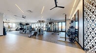 Located at 321 Main St in Hackensack, NJ, this expansive fitness center offers a generously sized, single-floor exercise space thoughtfully designed for a comprehensive workout experience. The facility features a clean, modern aesthetic with white walls, linear recessed lighting, and multiple ceiling fans that contribute to a bright, well-ventilated atmosphere throughout.

The space is divided into distinct functional zones. A dedicated cardio area lines the perimeter, equipped with treadmills, elliptical machines, stair climbers, stationary bikes, and rowing machines, all positioned to take advantage of large floor-to-ceiling windows that flood the room with natural light and offer open outdoor views. A strength training zone houses a robust selection of cable machines, multi-functional cable towers, and adjustable benches, complemented by a well-stocked free weights area featuring dumbbells, kettlebells, medicine balls, stability balls, and weight plates on vertical racks.

A separate functional training zone is outfitted with a dedicated storage rack for accessories including TRX suspension straps and resistance equipment. Full-length mirrors are strategically placed throughout, enhancing the sense of space. The flooring transitions between resilient rubber and smooth hardwood-style surfaces to suit different training needs. A storage credenza and decorative wall art add a polished, hospitality-inspired finish to the space.