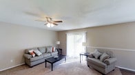 This single-floor space centers around a welcoming living room designed for both comfort and functionality. The room features wall-to-wall carpeting in a warm neutral tone, complemented by beige walls that create a calm, understated ambiance throughout. A ceiling fan with an integrated light fixture is mounted overhead, providing both air circulation and ambient lighting.

The living room is furnished with two grey upholstered sofas accented with decorative pillows in muted coral and blue tones, arranged around a dark wood coffee table. A pair of floor lamps flank the room, adding soft supplemental lighting. A notable feature is the sliding glass door dressed with vertical blinds, which serves as the room's primary source of natural light, flooding the space with brightness and offering a view of the outdoors.

A subtle chair rail detail runs along one wall, adding a modest architectural accent to the otherwise straightforward design. The overall layout is open and uncluttered, making efficient use of the available space while maintaining a comfortable, livable atmosphere suitable for everyday relaxation and casual entertaining.