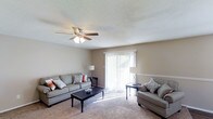 This single-floor space centers around a welcoming living room designed for both comfort and functionality. The room features wall-to-wall carpeting in a warm neutral tone, complemented by beige walls that create a calm, understated ambiance throughout. A ceiling fan with an integrated light fixture is mounted overhead, providing both air circulation and ambient lighting.

The living room is furnished with two grey upholstered sofas accented with decorative pillows in muted coral and blue tones, arranged around a dark wood coffee table. A pair of floor lamps flank the room, adding soft supplemental lighting. A notable feature is the sliding glass door dressed with vertical blinds, which serves as the room's primary source of natural light, flooding the space with brightness and offering a view of the outdoors.

A subtle chair rail detail runs along one wall, adding a modest architectural accent to the otherwise straightforward design. The overall layout is open and uncluttered, making efficient use of the available space while maintaining a comfortable, livable atmosphere suitable for everyday relaxation and casual entertaining.