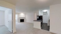 This single-floor space showcases an open-concept layout that connects the living area and kitchen in a cohesive, functional design. The living room is finished with plush, light-colored carpeting and centers around a tile-surround fireplace with a classic mantel, serving as a natural focal point. Neutral white walls and flush ceiling lighting throughout contribute to a clean, bright aesthetic.

The kitchen is positioned on one end of the open floor plan, delineated by a transition to dark hardwood-style flooring. It features white upper cabinetry, a dark countertop peninsula that provides additional workspace and a visual boundary between the two areas, and stainless steel appliances including a gas range and refrigerator. A built-in range hood is mounted beneath the upper cabinets, adding a practical touch. A full-height pantry or storage closet with bifold doors is accessible from the kitchen area.

Natural light filters in through a window visible in the background near the fireplace wall. The overall ambiance is clean, modern, and move-in ready, with a neutral palette that offers versatility for various interior styles.
