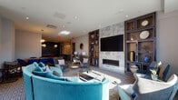 Located at 50 Lecount Place in New Rochelle, NY, this single-floor amenity space is designed to offer residents a sophisticated and versatile gathering environment. The main lounge area serves as the centerpiece, anchored by a striking built-in shelving unit with a textured stone accent wall, a large flat-screen TV, and a sleek linear fireplace. Plush teal curved sectional sofas and grey upholstered armchairs are arranged to encourage both social interaction and relaxed conversation, while recessed lighting and pendant fixtures create a warm, inviting ambiance. Natural light filters in through large windows and glass double doors that open to an outdoor terrace, enhancing the sense of openness throughout the space. A dedicated seating nook with a decorative bookcase offers a quieter retreat. The game and dining area features a pool table, communal tables, and bold red seating accented by stylish globe pendant lights, adding an energetic contrast to the lounge. A fully equipped kitchen anchors the entertaining side of the space, showcasing grey flat-front cabinetry, stainless steel appliances, a teal mosaic tile backsplash, light wood-look flooring, and a marble-topped island with bar seating — ideal for hosting and catered events. Throughout, the design palette of teal, grey, and warm wood tones creates a cohesive, contemporary aesthetic.