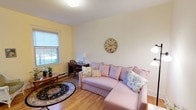This inviting single-floor space exudes a warm, cottage-inspired charm that is both comfortable and functional. The living area serves as the heart of the space, featuring rich hardwood floors that run throughout and contribute to the room's welcoming ambiance. Soft, creamy yellow walls complement the light, neutral ceiling, creating a bright and airy atmosphere enhanced by natural light streaming in through a classic double-hung window. A plush blush pink sectional sofa, dressed with decorative floral and striped throw pillows, anchors the seating area and adds a gentle, inviting touch. A traditional oval area rug in deep, rich tones grounds the space and adds a layer of visual interest underfoot. A vintage-style wall clock serves as a tasteful focal point on the main wall, while a wooden writing desk paired with a classic Windsor chair nestled near the window offers a quiet nook for work or study. A small side table with a floral arrangement and a table lamp adds warmth and a personal touch to the corner. The overall ambiance is relaxed and refined, blending timeless furnishings with soft, pastel hues to create a space that feels both lived-in and thoughtfully curated.