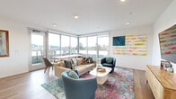 This well-appointed single-floor space offers a seamless blend of modern design and functional living. The open-plan layout integrates a living room and kitchenette into one cohesive area, complemented by a hallway that connects the space efficiently.

The living area is a standout, anchored by warm hardwood flooring and recessed lighting throughout. Floor-to-ceiling corner windows flood the room with natural light and frame expansive outdoor views of the surrounding landscape and cityscape beyond. A sliding glass door leads to an outdoor balcony, further extending the sense of openness. A wall-mounted flat-screen television, a media console, and a vibrant area rug complete the comfortable yet contemporary atmosphere.

The kitchenette is thoughtfully designed with light wood cabinetry, a white quartz countertop, a herringbone-tiled backsplash, and open floating shelves crafted from rich, dark-stained wood. A built-in beverage cooler and a single-basin sink add to the unit's practical functionality.

White walls throughout provide a clean, neutral backdrop that accentuates the colorful décor accents and artwork displayed in the space. The overall ambiance is bright, airy, and modern, making excellent use of its compact footprint while delivering a polished, inviting environment.