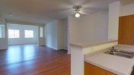 This single-floor residence offers a practical and comfortable layout designed for easy living. The open-concept floor plan creates a natural flow between the kitchen and living areas, making the space feel connected and spacious. Rich hardwood flooring extends throughout, adding warmth and visual continuity to the interior. A ceiling fan with a built-in light fixture is centrally positioned, providing both airflow and ambient lighting to the main living space. The kitchen features a peninsula-style counter with granite-look countertops and warm wood cabinetry, providing ample workspace and a natural divider between the kitchen and the rest of the living area. Neutral-toned walls throughout create a clean, versatile backdrop well-suited to a variety of furnishing styles. A window at the far end of the space allows natural light to filter in, brightening the interior and enhancing the open feel. A doorway visible in the background suggests additional rooms or storage beyond the main living space. Overall, the residence presents a straightforward, functional design with quality finishes that balance comfort and practicality.