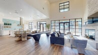 This thoughtfully designed single-floor community clubhouse offers an open, airy atmosphere anchored by soaring ceilings and an abundance of natural light streaming through expansive floor-to-ceiling black-framed windows and glass doors. Rich wood-look flooring flows throughout, grounding the contemporary aesthetic.

The main gathering area is generously proportioned, featuring a shuffleboard table as a central entertainment focal point, complemented by comfortable lounge seating with navy blue sofas, accent chairs, and a round dining area with woven rattan chairs. A stacked-stone fireplace with a mounted TV creates a cozy seating nook, adding warmth and texture to the space.

The fully equipped kitchen is highlighted by light wood cabinetry, a stainless steel refrigerator, a statement range hood, quartz countertops, and a distinctive teal hexagonal tile backsplash with open shelving for both function and style. A kitchen island provides additional prep and gathering space.

Near the entrance, a welcoming lounge with warm wood furnishings and a sculptural chandelier transitions into a dedicated workspace with a desk and shelving. Bold, oversized artwork adorns the walls throughout, reinforcing the space's modern, curated character. Large glass doors open directly to outdoor areas, seamlessly connecting the interior to the surrounding community.