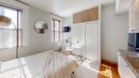 Located at 3 Cottage Ave in Quincy, Massachusetts, this efficiently designed one-bedroom, one-bathroom unit makes smart use of its intimate footprint. The open layout seamlessly integrates the sleeping area with a built-in kitchenette, which features light wood cabinetry, granite-style countertops, an undermount sink, a microwave, and a wall-mounted air conditioning unit — providing all essential amenities in a compact, organized arrangement. Flooring transitions between warm hardwood near the kitchenette and soft carpet in the sleeping area, adding texture and comfort throughout.

The bedroom area is bright and inviting, benefiting from multiple windows dressed with blinds that allow generous natural light to filter in. A closet with overhead storage cabinetry maximizes vertical space, while clean, light-gray walls contribute to an airy, calming atmosphere. A round wall mirror and framed artwork add thoughtful decorative touches.

The bathroom is well-appointed with classic white subway tile lining the tub-shower surround, accented by a bold navy tile border. A mosaic tile floor, wall-mounted towel warmer, and built-in amenities round out this functional yet stylish space. Overall, the unit offers a clean, modern aesthetic with practical features well-suited for efficient living in the Quincy area.