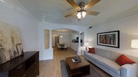 This single-floor residence showcases a thoughtfully designed open-concept layout that creates a natural flow between the main living spaces. The living room serves as the heart of the home, featuring a tray ceiling with a decorative ceiling fan, warm hardwood-style flooring, and ample space for comfortable seating. A tasteful area rug and wood coffee table anchor the space, complemented by a framed piece of wall art that adds a vibrant splash of color.

Elegant decorative columns and a graceful arched opening serve as a striking architectural transition into the dining area, which is illuminated by a pendant light fixture and furnished with a classic round dining table and upholstered chairs. This thoughtful design detail lends the space a refined, formal character without sacrificing openness.

Beyond the dining area, the kitchen is visible with dark countertops that contrast against white cabinetry, offering a clean and functional aesthetic. The consistent hardwood-style flooring throughout ties all areas together cohesively. Generous ceiling heights, neutral white walls, and an abundance of light contribute to a bright and airy ambiance, making this residence both welcoming and visually spacious.