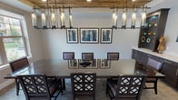 Located at 131 E Bardin Rd in Arlington, Texas, this well-appointed single-floor space serves as a sophisticated private dining and meeting room. The centerpiece of the main area is a large dark-wood dining table surrounded by upholstered chairs with decorative lattice-back detailing, comfortably seating a generous number of guests. Overhead, a striking rectangular pendant chandelier with cylindrical glass fixtures hangs beneath a distinctive reclaimed wood ceiling panel, adding warmth and rustic character to the otherwise contemporary interior. Recessed lighting complements the pendant fixtures throughout the space. Large windows along one wall flood the room with natural light and offer views of the surrounding outdoor greenery and courtyard, enhancing the open, welcoming atmosphere. A curated gallery of framed abstract artwork adorns the walls, contributing to the room's refined aesthetic. Along one side, a built-in dark cabinetry credenza with light stone countertops provides ample surface space, accompanied by an open display shelf and a wall-mounted flat-screen television — making this area ideal for presentations or casual gatherings. Neutral-toned carpet with subtle linear patterning grounds the space, while an adjacent area with hardwood-style flooring and additional windows extends the sense of openness.