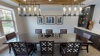 Located at 131 E Bardin Rd in Arlington, Texas, this well-appointed single-floor space serves as a sophisticated private dining and meeting room. The centerpiece of the main area is a large dark-wood dining table surrounded by upholstered chairs with decorative lattice-back detailing, comfortably seating a generous number of guests. Overhead, a striking rectangular pendant chandelier with cylindrical glass fixtures hangs beneath a distinctive reclaimed wood ceiling panel, adding warmth and rustic character to the otherwise contemporary interior. Recessed lighting complements the pendant fixtures throughout the space. Large windows along one wall flood the room with natural light and offer views of the surrounding outdoor greenery and courtyard, enhancing the open, welcoming atmosphere. A curated gallery of framed abstract artwork adorns the walls, contributing to the room's refined aesthetic. Along one side, a built-in dark cabinetry credenza with light stone countertops provides ample surface space, accompanied by an open display shelf and a wall-mounted flat-screen television — making this area ideal for presentations or casual gatherings. Neutral-toned carpet with subtle linear patterning grounds the space, while an adjacent area with hardwood-style flooring and additional windows extends the sense of openness.