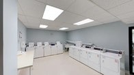This single-floor communal laundry facility is designed with practicality and efficiency in mind. The room features a generous arrangement of top-load washing machines lined along one wall and a matching set of front-load dryers positioned along the adjacent wall, providing ample capacity for multiple users simultaneously. The space is finished with smooth, light-colored walls painted in a soft blue-gray tone, lending a clean and calm atmosphere. Overhead, a drop ceiling with recessed fluorescent panel lighting ensures the room is evenly and brightly illuminated throughout. The flooring is a light-toned vinyl surface that is easy to maintain and contributes to the overall sense of cleanliness. A utility sink is visible in the corner, offering added convenience for pre-treating garments or other laundry needs. A ventilation duct runs along the upper portion of the back wall, indicating proper airflow management within the facility. The layout is open and uncluttered, allowing residents to move freely between machines. Overall, the space is thoughtfully organized to support a high-functioning, shared laundry environment.