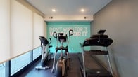 This fitness center is a thoughtfully designed single-level exercise space that maximizes functionality within a compact footprint. The room is anchored by a striking teal accent wall emblazoned with the motivational phrase "Don't Wish For It, Work For It," creating an energizing focal point that sets the tone for the entire space. Recessed ceiling lighting provides consistent illumination throughout, while large windows fitted with roller shades allow natural light to filter in and offer views to the exterior.

The flooring consists of durable speckled rubber gym matting, practical for both safety and equipment placement. Full-length mirrors line one wall, visually expanding the space and allowing users to monitor their form during workouts. The cardio zone features treadmills and elliptical machines positioned to face the motivational wall, while the strength training area is stocked with a tiered dumbbell rack, kettlebells, a weight bench, and a barbell setup. Additional equipment includes a medicine ball tower, foam rollers, yoga mats, and resistance bands organized neatly on wall-mounted storage racks. A wall-mounted television, sanitation dispensers, and waste bins round out the practical amenities, making this a fully self-contained fitness facility.