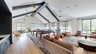 Located at 1013 Hunters Glen Dr in Plainsboro, NJ, this single-floor amenity space offers a well-appointed community lounge designed with contemporary style and multiple distinct functional areas. The game room is the architectural centerpiece, boasting dramatic vaulted ceilings accented by bold black exposed beams, warm pendant lighting, and an abundance of natural light streaming through large black-framed windows. The space accommodates pool tables, a foosball table, and generous seating, all set atop light hardwood flooring. Adjacent to the game room, a sophisticated lounge area features a full-height large-format tile accent wall housing both a wall-mounted TV and a sleek linear gas fireplace, flanked by leather armchairs and a sectional sofa for comfortable gathering. A separate bar and kitchen area showcases a warm wood tray ceiling, a long white quartz countertop with bar seating, dark cabinetry, a utility sink, and dual wall-mounted TVs — ideal for social events. Throughout the space, floor-to-ceiling glass partitions and black steel-framed windows maintain visual connectivity while defining zones. Bistro-style high-top tables near the windows provide additional casual seating with pleasant outdoor views, completing this well-rounded, amenity-rich community space.