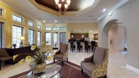 This sophisticated single-floor space exudes warmth and elegance through its thoughtfully designed interior. The main lounge area is anchored by a dramatic tray ceiling finished in a deep, contrasting tone, adorned with an ornate wrought-iron chandelier that serves as a striking focal point. Crown molding frames the room, adding a refined architectural detail throughout.

The seating area features plush upholstered armchairs with decorative floral patterning, arranged around a glass-topped coffee table and accented by vibrant yellow floral arrangements that complement the warm, golden-toned walls. Large windows along the exterior wall flood the space with abundant natural light and offer views of the outdoor surroundings.

Transitioning seamlessly from the lounge, the kitchen and bar area is outfitted with dark cabinetry, stainless steel appliances, and pendant lighting above a generous bar counter lined with high-back bar stools. An arched architectural detail frames the passage between areas, lending a Mediterranean-inspired character to the overall design.

Light tile flooring extends throughout, contrasted by a richly patterned area rug in the lounge zone. The overall ambiance is inviting and polished, making this a well-appointed gathering or community space designed for both comfort and style.