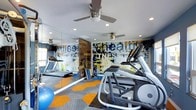 This single-floor fitness center is designed to inspire and energize with a bold, contemporary aesthetic. The space is immediately defined by a striking, large-scale graphic mural spanning a full wall, featuring dynamic typography with words like "health" and "fitness" rendered in an urban, expressive style with splashes of blue, yellow, and white. Complementing the mural, a full-length mirror wall visually expands the room and allows users to monitor their form during workouts.

The flooring is a standout design element, featuring a geometric patchwork pattern in contrasting shades of orange, blue, and grey carpet tiles that add energy and visual interest underfoot. Recessed ceiling lighting provides bright, even illumination throughout, while multiple ceiling fans ensure comfortable air circulation during intense workouts.

The fitness floor is equipped with quality cardio machines, including treadmills and an elliptical trainer from the Matrix brand, as well as free weights and exercise accessories such as a stability ball. A wood-paneled accent wall adds a warm, textural contrast to the otherwise modern and industrial-inspired interior. The overall atmosphere is motivating, functional, and thoughtfully designed to support a comprehensive fitness experience.