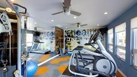 This single-floor fitness center is designed to inspire and energize with a bold, contemporary aesthetic. The space is immediately defined by a striking, large-scale graphic mural spanning a full wall, featuring dynamic typography with words like "health" and "fitness" rendered in an urban, expressive style with splashes of blue, yellow, and white. Complementing the mural, a full-length mirror wall visually expands the room and allows users to monitor their form during workouts.

The flooring is a standout design element, featuring a geometric patchwork pattern in contrasting shades of orange, blue, and grey carpet tiles that add energy and visual interest underfoot. Recessed ceiling lighting provides bright, even illumination throughout, while multiple ceiling fans ensure comfortable air circulation during intense workouts.

The fitness floor is equipped with quality cardio machines, including treadmills and an elliptical trainer from the Matrix brand, as well as free weights and exercise accessories such as a stability ball. A wood-paneled accent wall adds a warm, textural contrast to the otherwise modern and industrial-inspired interior. The overall atmosphere is motivating, functional, and thoughtfully designed to support a comprehensive fitness experience.