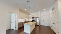 This contemporary single-floor residence showcases a thoughtfully designed open-concept layout with high-end finishes throughout. The kitchen serves as the heart of the home, centered around a white quartz island beneath a trio of sleek pendant lights. Two-toned cabinetry—white upper cabinets and warm wood-grain lowers with matte black hardware—frames a white subway tile backsplash. A full suite of stainless steel appliances, including a refrigerator, range, microwave, and dishwasher, complete the space. Dark hardwood flooring flows seamlessly throughout the residence, providing a rich contrast to the crisp white walls.

The living area is bright and open, benefiting from natural light and direct access to a private balcony through a glass-panel door. The primary bedroom is generously proportioned, featuring a ceiling fan and direct access to an en-suite bathroom and a walk-in closet equipped with wire shelving systems. The primary bath offers a dual-sink vanity with quartz countertops, a glass-enclosed walk-in shower with white tile surround, and black fixture accents. A secondary bathroom mirrors the modern aesthetic with a dual-sink vanity, large mirror, and black hardware. The overall ambiance is clean, minimal, and move-in ready.