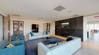 This thoughtfully designed single-floor amenity space offers a diverse range of areas crafted for both relaxation and entertainment. The main lounge is an inviting focal point, featuring light-toned modular sofas arranged around a sleek linear fireplace set against a dramatic dark stone accent wall, with a large wall-mounted display and recessed lighting overhead. Light hardwood flooring flows throughout the open-plan areas, lending warmth and continuity to the space.

The fully equipped kitchen is a standout feature, anchored by a generous marble-topped island with built-in induction cooking and wrapped in an striking geometric metal pendant fixture. Warm wood cabinetry, dark tile backsplash, and stainless steel appliances complete the professional-grade setup. A casual dining area nearby is bathed in natural light from floor-to-ceiling corner windows, offering sweeping urban views of the surrounding cityscape.

A secondary lounge zone provides flexible seating with frosted glass partitions for privacy, while a window-lined nook with striped bench seating frames scenic outdoor views. The game room accommodates a full billiards table alongside additional recreational furnishings. A dedicated screening room, finished in dark patterned wall panels with ambient wall sconces and generously arranged lounge chairs, rounds out this impressive amenity floor.