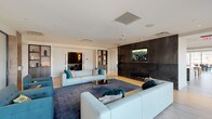 This thoughtfully designed single-floor amenity space offers a diverse range of areas crafted for both relaxation and entertainment. The main lounge is an inviting focal point, featuring light-toned modular sofas arranged around a sleek linear fireplace set against a dramatic dark stone accent wall, with a large wall-mounted display and recessed lighting overhead. Light hardwood flooring flows throughout the open-plan areas, lending warmth and continuity to the space.

The fully equipped kitchen is a standout feature, anchored by a generous marble-topped island with built-in induction cooking and wrapped in an striking geometric metal pendant fixture. Warm wood cabinetry, dark tile backsplash, and stainless steel appliances complete the professional-grade setup. A casual dining area nearby is bathed in natural light from floor-to-ceiling corner windows, offering sweeping urban views of the surrounding cityscape.

A secondary lounge zone provides flexible seating with frosted glass partitions for privacy, while a window-lined nook with striped bench seating frames scenic outdoor views. The game room accommodates a full billiards table alongside additional recreational furnishings. A dedicated screening room, finished in dark patterned wall panels with ambient wall sconces and generously arranged lounge chairs, rounds out this impressive amenity floor.