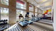 Located at 4650 N Central Ave in Phoenix, Arizona, this two-story fitness center — branded "Elevation" — offers a well-appointed and thoughtfully designed workout facility. The main level is anchored by soaring double-height ceilings and floor-to-ceiling windows framed in warm natural stone, flooding the space with abundant natural light and offering views of the surrounding palm tree-lined streetscape. A row of treadmills and elliptical trainers lines the window wall, complemented by a comprehensive free weight section featuring multiple dumbbell racks and adjustable benches. A wall of mirrors expands the visual space, while a bold striped carpet grounds the open floor plan. Strength and cable machines are distributed throughout, providing a full range of resistance training options. A staircase with warm wood-toned metal railings connects to the second floor, where additional cardio and strength equipment is arranged alongside the upper-level overlook. A dedicated functional training room on the lower level features stability balls and medicine balls set against vibrant accent walls in orange and red. Ceiling fans and recessed lighting ensure comfort throughout. Colorful wall art adds an energetic, motivating atmosphere to the entire facility.
