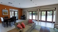 This single-story communal space serves as a welcoming lounge and activity area, thoughtfully designed to blend comfort with functionality. The interior features a neutral color palette accented by a warm terracotta-orange feature wall, creating an inviting and energetic atmosphere. Recessed lighting in the white ceiling provides consistent illumination throughout the space.

The lounge area is anchored by a plush taupe sofa adorned with vibrant coral and patterned throw pillows, positioned atop a striped area rug over light-toned tile flooring. A dark coffee table completes the seating arrangement, offering a casual gathering spot. Adjacent to the lounge, a dedicated workstation with a desk and ergonomic chair provides a practical workspace.

Large glass-paneled French doors line one wall, flooding the interior with natural light and offering direct views and access to an outdoor pool area surrounded by lush greenery. The seamless indoor-outdoor connection is a defining feature of this space, making it ideal for social gatherings. Bar-height seating near the windows further enhances the versatility of the layout, accommodating both relaxed lounging and more active use.