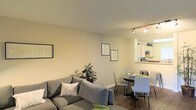 This single-floor space showcases an open-concept design that seamlessly connects the living, dining, and kitchen areas into one cohesive environment. The living area is anchored by a comfortable gray sectional sofa, accented with decorative pillows and complemented by a lush indoor plant that adds a touch of greenery. Framed artwork on the warm, neutral-toned walls adds a curated, welcoming feel to the space.

Adjacent to the living area, a compact dining space accommodates a round table with modern chairs, making it ideal for intimate meals. Track lighting overhead provides focused illumination, enhancing the functionality of the combined space.

The kitchen is visible through a pass-through opening, featuring light wood cabinetry and standard appliances. This design element maintains an open, airy connection between the cooking and living spaces while defining each area distinctly.

Dark wood-style flooring runs throughout, providing a grounding contrast to the light-colored walls and ceiling. Overall, the space maintains a clean, minimalist aesthetic with practical furnishings and a layout that maximizes the available footprint efficiently.