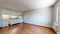 Located at 36 West St in Akron, Ohio, this single-floor unit offers a straightforward and functional layout finished with warm wood-look flooring that extends consistently throughout the space. The open living area provides generous room to arrange furnishings, with neutral light gray walls and clean white ceilings creating a bright, airy atmosphere. Baseboard heating runs along the perimeter of the main living areas, ensuring efficient climate control throughout the colder months.

The kitchen is positioned along one wall and features white cabinetry both above and below the counter, a dark granite-look countertop, a white refrigerator, a white range, an under-cabinet light, and a tile backsplash — offering a compact yet complete cooking space. A dedicated hallway leads to a separate room, providing privacy and separation from the main living area.

The bedroom includes a wall-mounted air conditioning unit for independent climate control, a window with views of the surrounding neighborhood streetscape, and a sliding closet door offering practical storage. Throughout the unit, the clean, neutral palette and consistent flooring make it easy to personalize the space. This unit presents a practical and comfortable living environment in the heart of Akron.