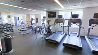 This fitness center offers a comprehensive workout environment designed to accommodate a variety of exercise routines. The space is outfitted with a full suite of modern cardio equipment, including multiple treadmills, elliptical trainers, stationary bikes, and rowing machines, all arranged to provide ample room for movement and easy navigation between stations. Several of the machines feature individual built-in monitors, allowing users to track their performance metrics or enjoy personal entertainment during workouts. Wall-mounted flat-screen televisions are positioned at eye level throughout the space, ensuring visibility from multiple workout stations. Large windows line the walls, flooding the room with natural light and providing views of the surrounding cityscape, creating an energizing atmosphere. The flooring consists of dark, durable rubberized material well-suited for high-traffic fitness use, while the drop ceiling with recessed linear lighting contributes to a clean, modern aesthetic. The overall layout is open and well-organized, offering a functional and motivating environment suitable for both casual exercisers and dedicated fitness enthusiasts.