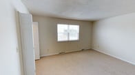 This single-floor space presents a clean and functional layout designed for comfortable everyday living. The interior is finished with wall-to-wall beige carpeting that adds warmth underfoot and contributes to a neutral, inviting atmosphere throughout. The walls are painted in a soft, light beige tone, creating a calm and cohesive look that complements the overall simplicity of the space. A prominent window along the main wall allows natural light to fill the room, brightening the interior and offering a view to the outdoors. The ceiling features a classic textured finish, adding subtle depth and character to the space. A white paneled closet door is visible along one wall, providing convenient built-in storage. Floor-level air vents are positioned along the baseboards, indicating a central HVAC system for year-round climate comfort. White trim and baseboards frame the room neatly, providing a crisp contrast to the soft wall color. The room feels open and well-proportioned, with ample floor space that offers versatility for a variety of uses. Overall, the space maintains a tidy, move-in-ready condition with a straightforward and practical aesthetic.
