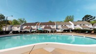This community amenity space offers two distinct areas designed for recreation and wellness. The centerpiece is a generously sized outdoor swimming pool with clear, blue-tinted water and a smooth surrounding deck ideal for lounging. The pool area is enclosed by a wrought iron fence and is framed by lush mature trees and a backdrop of multi-story residential buildings with classic white siding and brown rooflines. Poolside seating and shaded umbrella tables provide comfortable spaces to relax under open skies.

Adjacent to the outdoor area, the fitness center is a compact yet functional space with a purposeful layout. The room is finished with carpeted flooring and features bold dark accent walls that contrast with crisp white upper walls. High clerestory windows allow natural light to filter in, lending an airy feel despite the room's focused footprint. The space is equipped with a multi-station cable weight machine, a stationary bike, and additional exercise equipment suited for a full-body workout. A ceiling fan and pendant lighting keep the space comfortable, while built-in shelving and a large sliding glass door with views of the surrounding greenery add practical and aesthetic appeal.