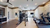 Located at 3000 Dominion Ridge Circle in Morrisville, NC, this single-floor fitness center is thoughtfully designed to support a full range of workout routines. The space is organized into distinct functional zones, creating a structured yet open environment for exercise.

The main workout area is generously sized and filled with natural light streaming through large windows fitted with horizontal blinds. Multiple ceiling fans throughout the space ensure comfortable air circulation during workouts. Recessed lighting provides consistent, even illumination across all areas.

The cardio zone lines the windowed perimeter and is equipped with a row of treadmills, elliptical trainers, and stair climbers, all oriented to take advantage of the natural light. A dedicated spin studio features several stationary cycling bikes and is enclosed with glass partitions and floor-to-ceiling mirrors, contributing to an open, airy feel.

The strength training area is anchored by a professional-grade cable machine system and a power rack with an attached bench, positioned on a dark rubberized floor mat. A mirrored wall and wall-mounted flat-screen TVs enhance the space throughout.

A wood-paneled accent wall adds a warm, contemporary design element, complemented by a built-in cubby storage unit. French doors provide access between sections of the facility, reinforcing the space's clean, upscale aesthetic.